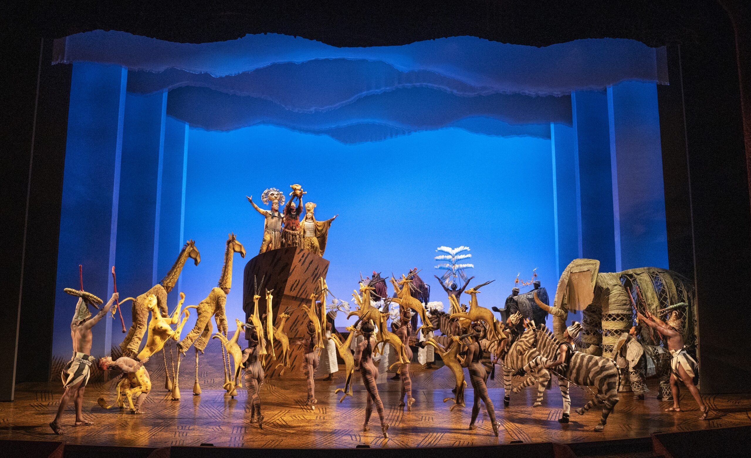 The company of "The Lion King" in front of a blue background. Performers are in detailed costumes to portray lions, giraffes, gazelles and other animals who live in the African savanna.
