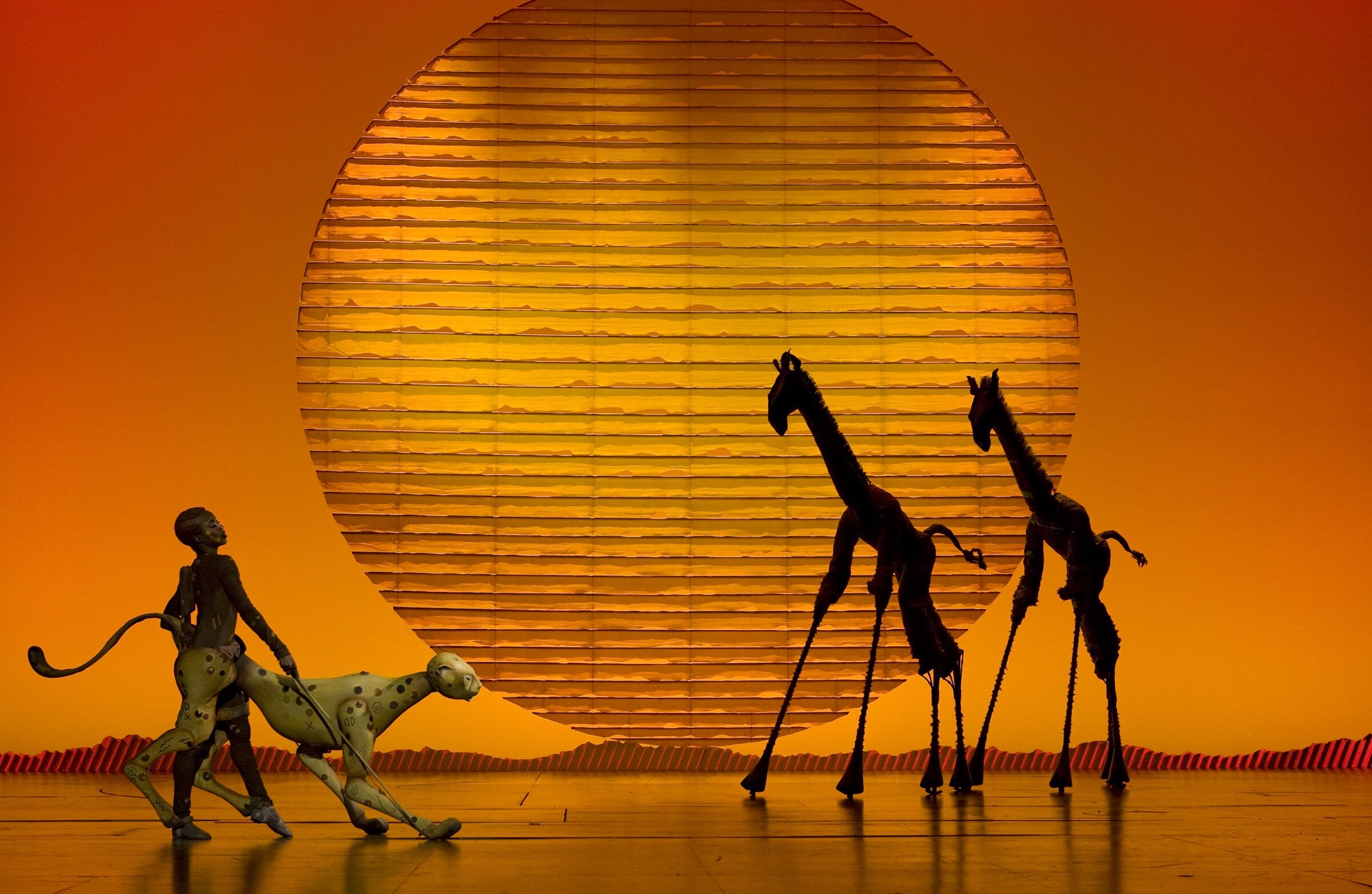 Actors in "The Lion King" perform on stage. To the left, an actor/puppeteer is a cheetah and to the right, actors/puppeteers portray two giraffes.
