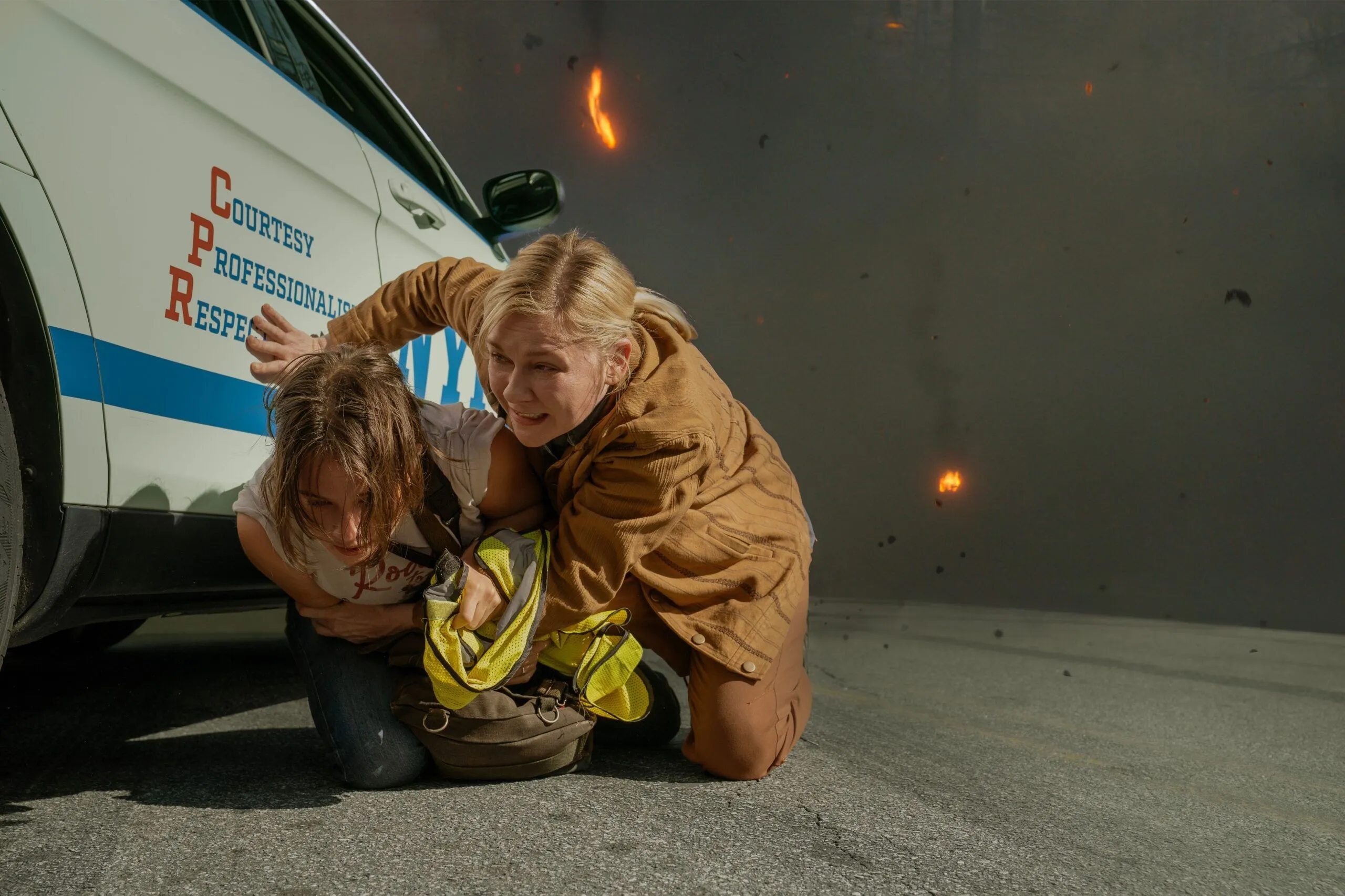 Actors Wagner Moura and Kirsten Dunst in a still image from the 2024 film, "Civil War." They are crouched beside a white and blue NYPD police car.
