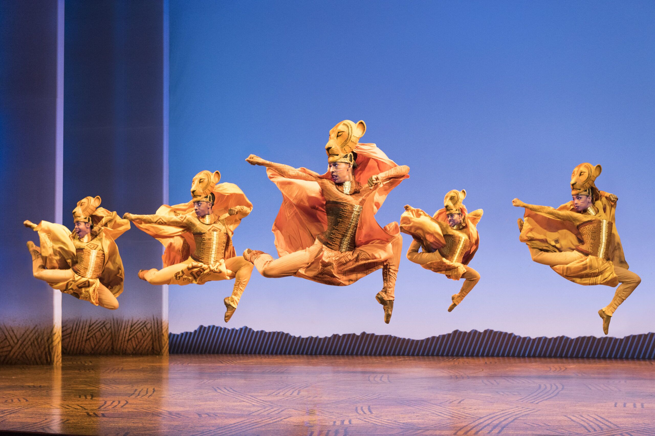 Several performers portraying the lionesses in "The Lion King" stage play leap in front of a blue background.