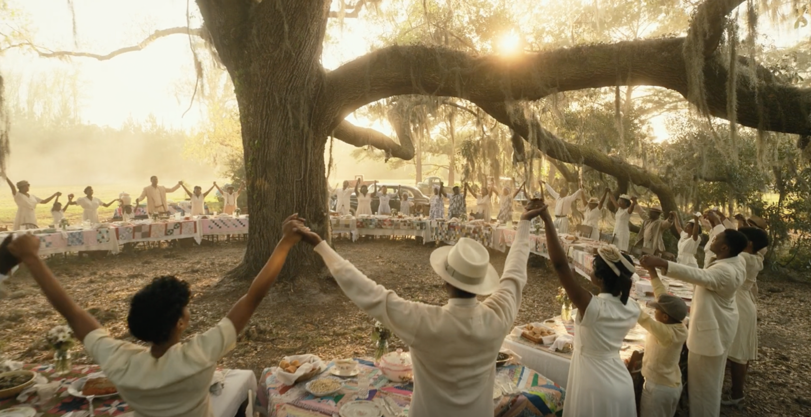 A still from the final scene of 2023's "The Color Purple." The majority of the cast are standing around tables that circle a tree, hands clasped and arms raised.