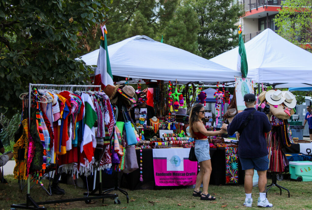 A photo of Cactus Girl Boutique's outdoor display at OYE Fest 2024. Vendors and shoppers stand in front of tents and clothing racks with colorful clothing.