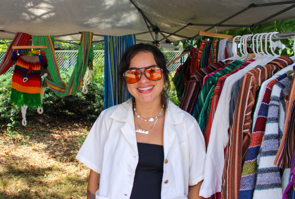 A photo of a person in a white button down tee in front of a rack of colorful clothing in an outdoor tent at a music festival.