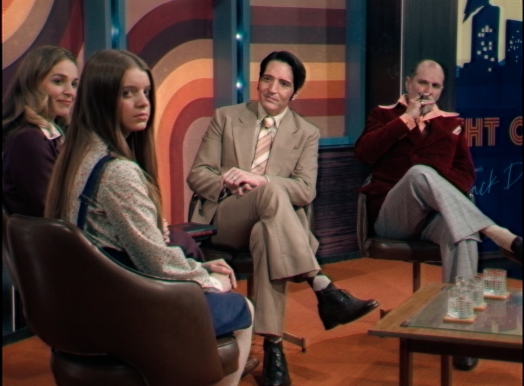 A still image from the IFC film, "Late Night with the Devil" that depicts a possessed teen girl, Lily (Ingrid Torelli), staring directly into the camera while on a colorful 1970s late night show set.